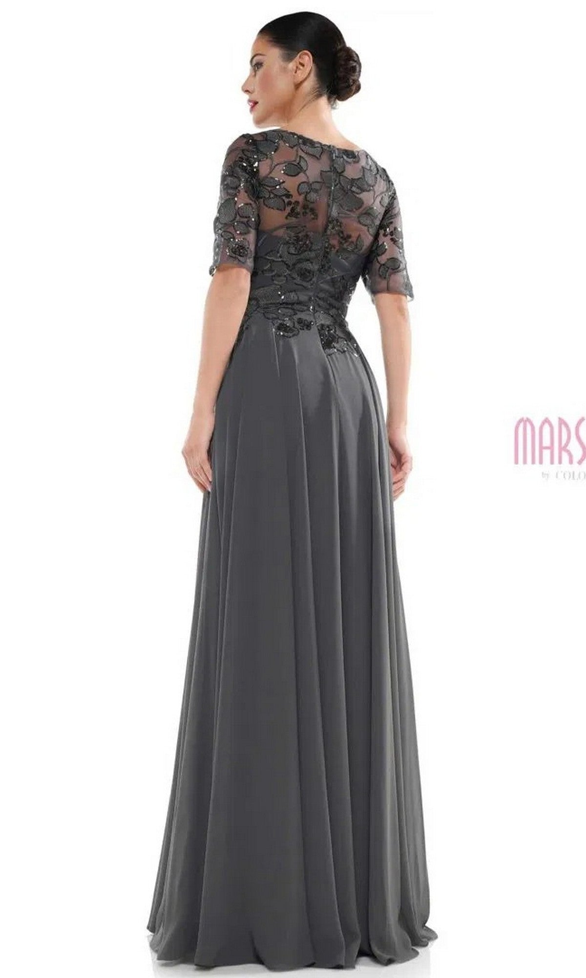 A woman stands facing away in the Marsoni M286 long formal dress, featuring a floor-length dark gray skirt and a sheer, embroidered bodice with half sleeves.